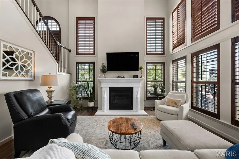 Two-story living area with a built-in fireplace, multiple windows with dark wood plantation shutters, a staircase with dark wood handrails, and wood-finish flooring — 2700 Grover Crossing Drive, Wildwood, MO 63040