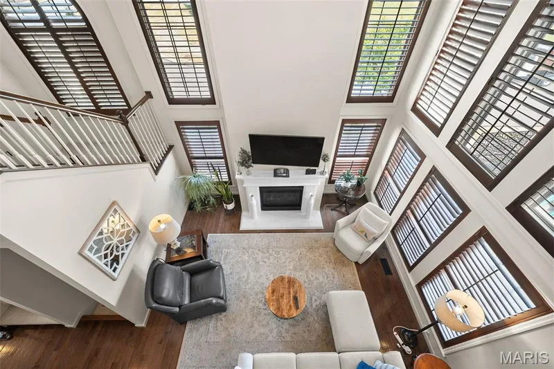 Two-story living area with fireplace, multiple windows with plantation shutters, wood-finish flooring, and a white staircase with wood handrail — 2700 Grover Crossing Drive, Wildwood, MO 63040