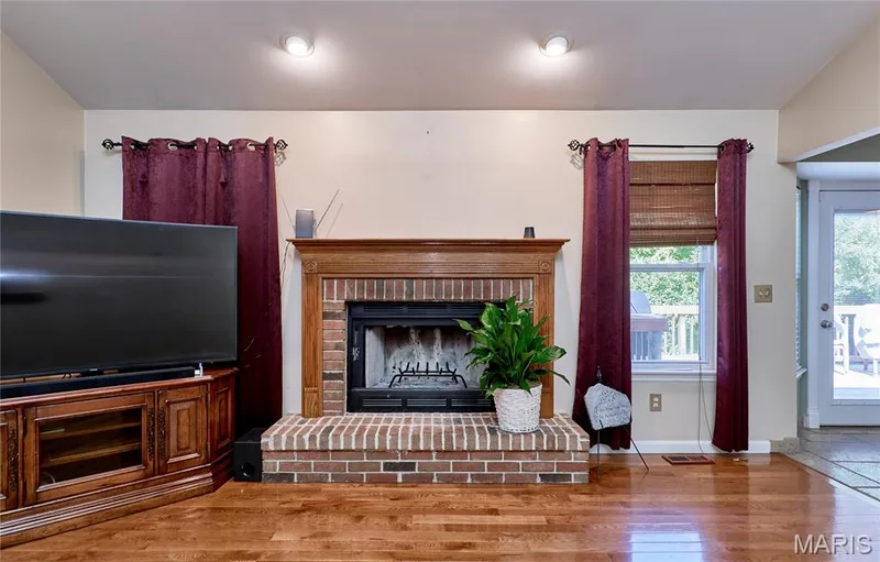 Spacious living area featuring hardwood flooring, a wood-trimmed fireplace with brick surround and gas insert, built-in wooden cabinetry with shelving, recessed ceiling lighting, and glass door access to outdoor space — 3641 Wind Chime Lane, St Charles, MO 63301