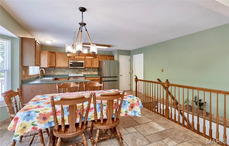 Open-concept kitchen and dining area featuring wood cabinetry, stainless steel appliances, tile flooring, a pendant light fixture, and direct access to an adjoining room with wood railings and soft green wall finishes — 3641 Wind Chime Lane, St Charles, MO 63301