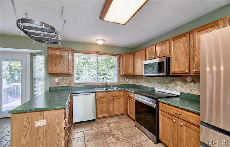 Spacious kitchen featuring warm wood cabinetry, green tile countertops, stainless steel appliances including a built-in microwave and dishwasher, ceramic tile flooring, and abundant natural light from multiple windows — 3641 Wind Chime Lane, St Charles, MO 63301