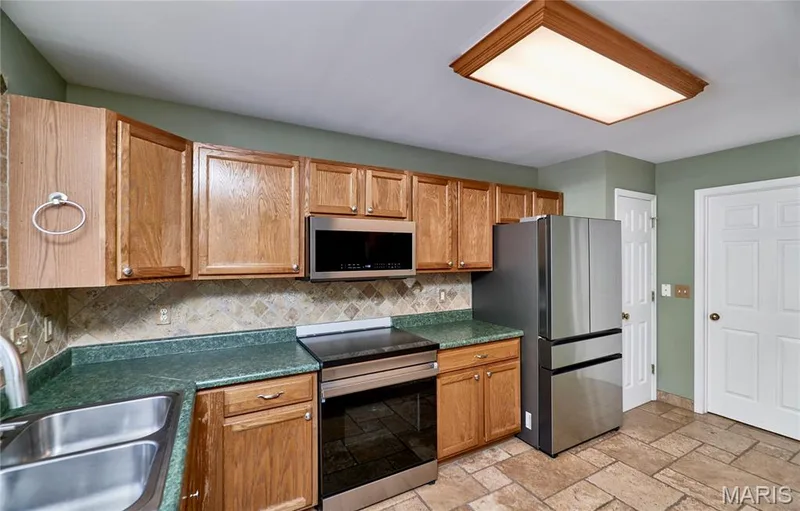 Galley kitchen featuring wood-finish cabinetry with hardware pulls, green-toned tile backsplash, green quartz countertops, stainless steel appliances including a built-in microwave and electric range, tile flooring, and recessed ceiling lighting — 3641 Wind Chime Lane, St Charles, MO 63301