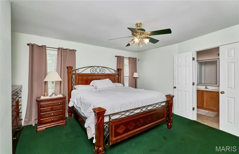 Spacious primary bedroom with wood-finish flooring, ceiling fan with integrated lighting, ornamental metal bed frame, wood nightstand with table lamp, and dual windows dressed in coordinating drapery — 3641 Wind Chime Lane, St Charles, MO 63301
