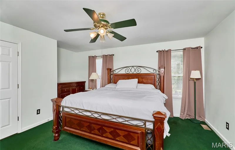 Spacious primary bedroom featuring a decorative metal headboard bed frame, ceiling fan with brass fixtures, dual windows dressed in coordinating drapery with plantation shutters, hardwood flooring, and soft recessed lighting throughout — 3641 Wind Chime Lane, St Charles, MO 63301