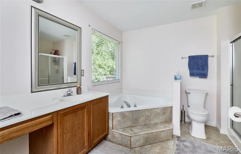 Master bathroom featuring a wood-finish vanity with dual sinks and ample counter space, a soaking tub with tile surround, a separate toilet enclosure, large framed mirror, and natural light from a window — 3641 Wind Chime Lane, St Charles, MO 63301