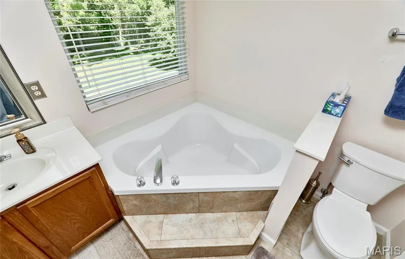 Corner soaking tub with tile surround, single vanity with integrated sink, toilet, and window with blinds — 3641 Wind Chime Lane, St Charles, MO 63301