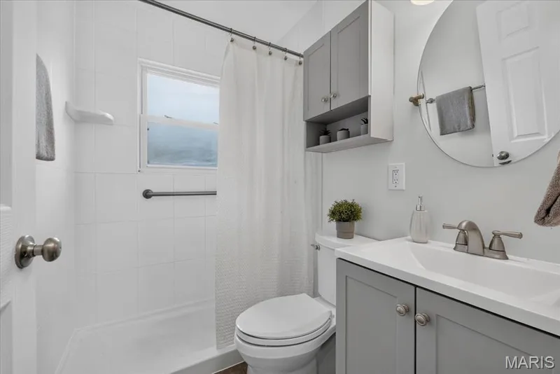 White tiled shower surround, built-in window, grab bar, gray vanity with integrated sink, and round mirror — 11 Toelle Drive, St Charles, MO 63304