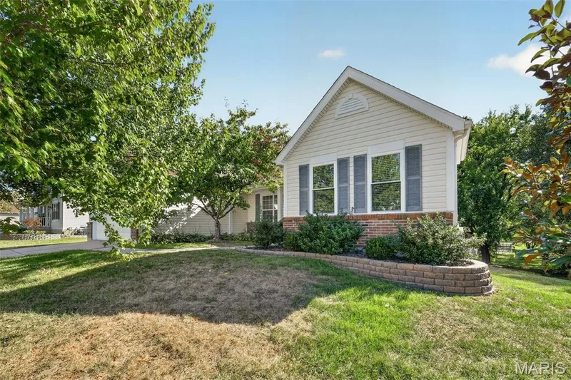 Gable roofline with dentil molding detail, light-colored siding, brick foundation, three windows with louvered shutters, and a block retaining wall — 155 Snake River Drive, Dardenne Prairie, MO 63368