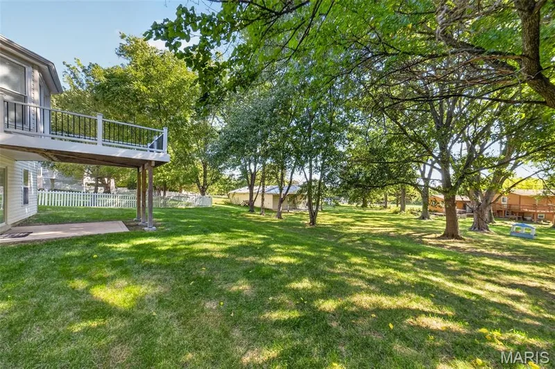 Expansive green lawn with mature trees, a raised deck with dark railings, a ground-level concrete patio, and white picket fencing — 155 Snake River Drive, Dardenne Prairie, MO 63368