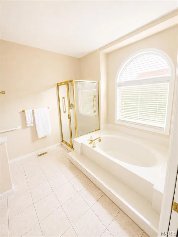 Spa-inspired primary bathroom featuring a soaking tub with integrated surround, frameless glass-enclosed shower with brushed gold hardware, ceramic tile flooring, and an arched transom window with plantation shutters providing natural light and ventilation — 1605 Ridge Bend Drive, Glencoe, MO 63038