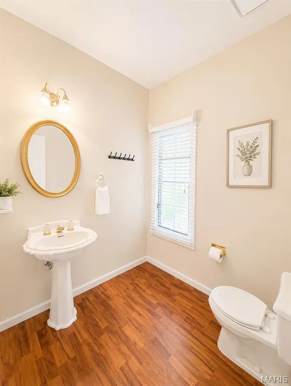 Elegant powder room featuring a classic pedestal sink with chrome fixtures, warm wood-finish flooring, soft neutral wall finishes, and an oval framed mirror — 1605 Ridge Bend Drive, Glencoe, MO 63038