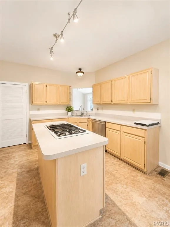 Spacious kitchen featuring natural wood cabinetry, a large center island with white countertop and built-in gas cooktop, stainless steel sink with window sill placement, and cork-finish flooring throughout — 1605 Ridge Bend Drive, Glencoe, MO 63038