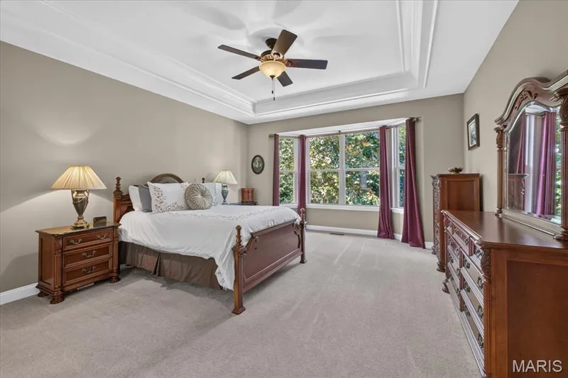 Primary suite w/recessed coffered ceiling and crown molding, ceiling fan with integrated lighting, panoramic bay window, neutral wall paint, and light-colored carpeting — 1307 Homestead Heights Drive, Wildwood, MO 63005