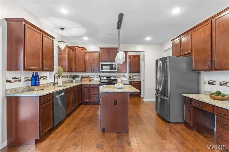 Spacious kitchen featuring wood-finish flooring, wood cabinetry, stone countertops, stainless steel appliances, and an island with pendant lighting — 3222 Starkville Street, St Charles, MO 63301