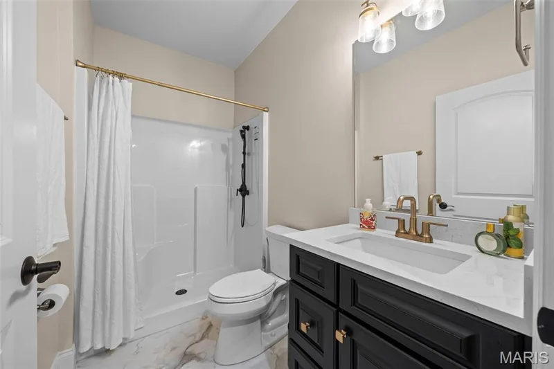 White shower-tub combination with black fixtures, a single vanity with a white countertop and gold-finish faucet, black cabinetry with gold hardware, and patterned floor tile — 193 Santa Elena Drive, Wentzville, MO 63385