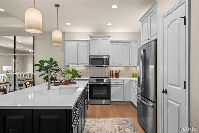 Kitchen featuring a large island with an integrated sink, light-toned upper cabinetry, stainless steel appliances, a patterned tile backsplash, and wood-finish flooring — 193 Santa Elena Drive, Wentzville, MO 63385