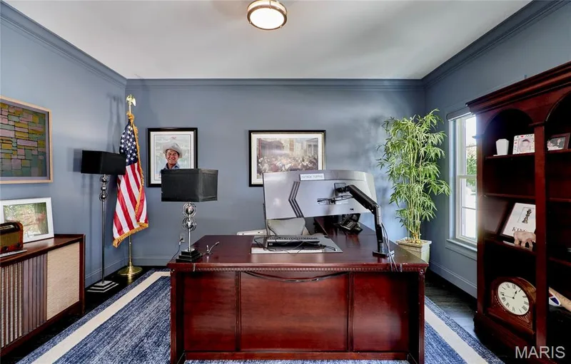 Executive home office featuring rich wood cabinetry and desk, recessed ceiling lighting, blue-toned walls, built-in shelving with glass doors, and a framed architectural print collection — 264 Greenshire Lane, Dardenne Prairie, MO 63368