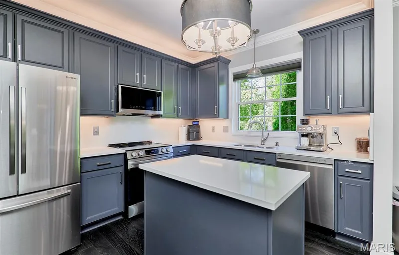Chef's kitchen featuring custom cabinetry in soft blue-gray finish, stainless steel appliances including a gas range and refrigerator, white quartz waterfall-edge island with ample seating, pendant lighting, and abundant natural light from large window overlooking landscaped grounds — 264 Greenshire Lane, Dardenne Prairie, MO 63368