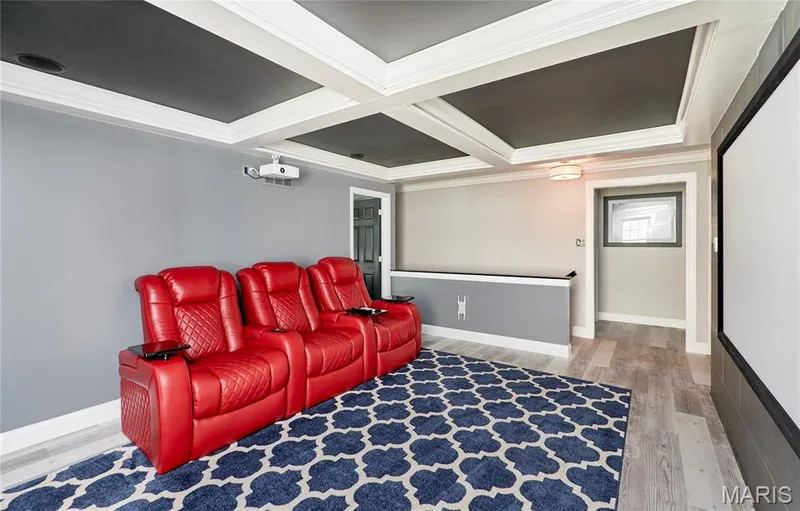 Dedicated media room featuring coffered ceiling with recessed lighting, built-in shelving with horizontal accent trim, wood-finish flooring, and geometric patterned area rug — 264 Greenshire Lane, Dardenne Prairie, MO 63368