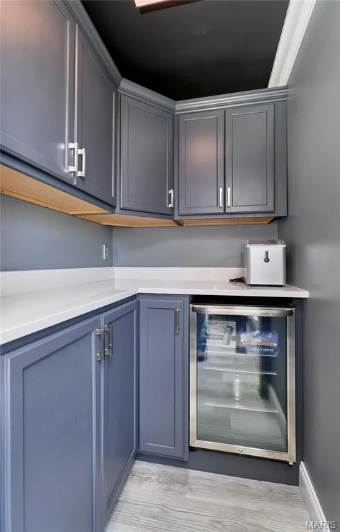Modern wet bar featuring gray cabinetry with chrome hardware, white quartz countertops, wood-finish upper shelving, beverage cooler with glass door, and neutral tile flooring throughout — 264 Greenshire Lane, Dardenne Prairie, MO 63368