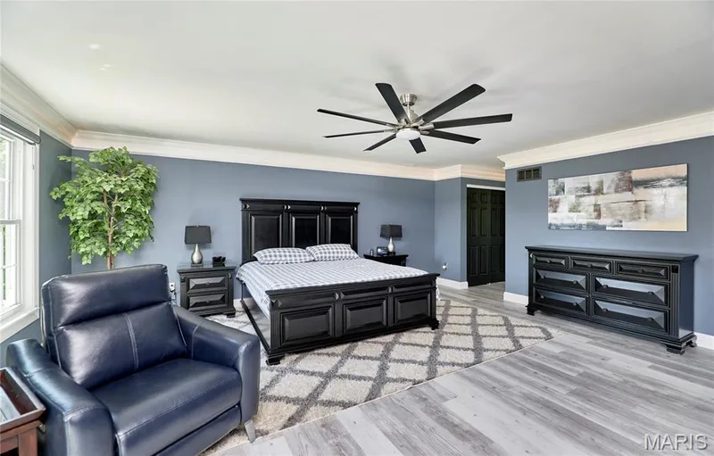 Spacious primary bedroom featuring a wood-finish plank floor, black wood bed frame with upholstered headboard, dual nightstands with task lighting, and a ceiling fan with brushed metal finish — 264 Greenshire Lane, Dardenne Prairie, MO 63368