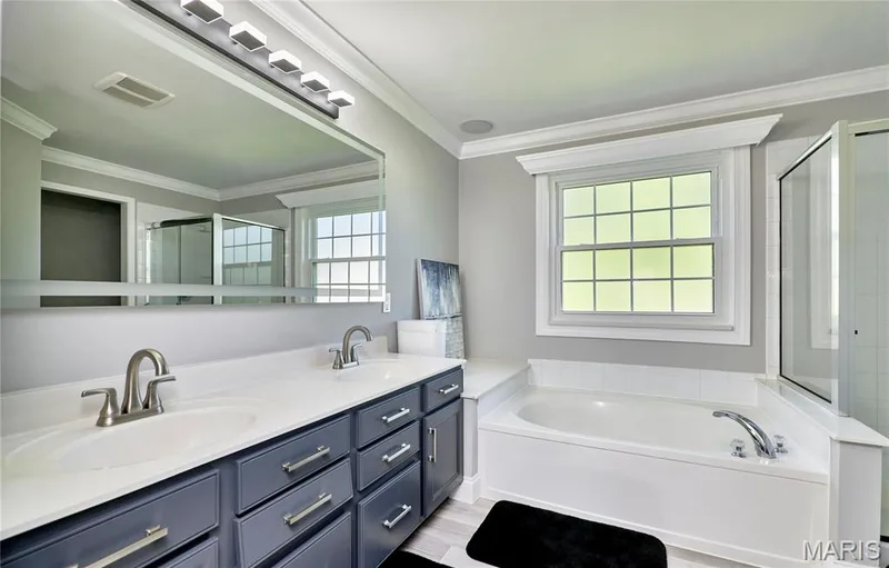 Spa-inspired primary bathroom featuring dual vanities with navy cabinetry and white quartz countertops, polished nickel faucets, large soaking tub with deck-mounted fixtures, extensive mirror wall with integrated lighting, and abundant natural light from grid-pattern windows — 264 Greenshire Lane, Dardenne Prairie, MO 63368