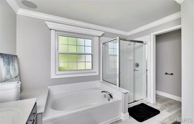 Spa-inspired primary bathroom featuring a soaking tub with chrome fixtures, a frameless glass-enclosed shower with tile surround, tile flooring, and abundant natural light from a large multi-pane window — 264 Greenshire Lane, Dardenne Prairie, MO 63368