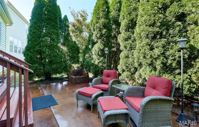 Private outdoor retreat featuring a spacious stamped concrete patio, wicker seating with cushioned frames, mature evergreen privacy hedging, integrated landscape lighting, and a multi-tiered hardscape design ideal for entertaining and relaxation — 264 Greenshire Lane, Dardenne Prairie, MO 63368
