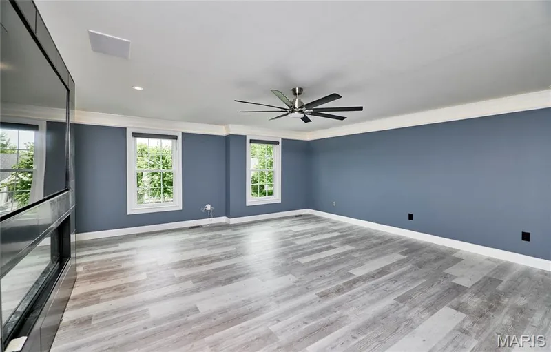 Spacious room featuring wood-finish flooring, two large windows, a built-in electric fireplace, a ceiling fan, and crown molding — 264 Greenshire Lane, Dardenne Prairie, MO 63368