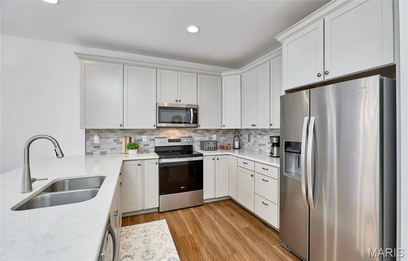 Kitchen featuring white cabinetry, stainless steel appliances, a double basin sink with a gooseneck faucet, light-toned countertops, and wood-finish flooring — 305 Copperfield Court, St Charles, MO 63301