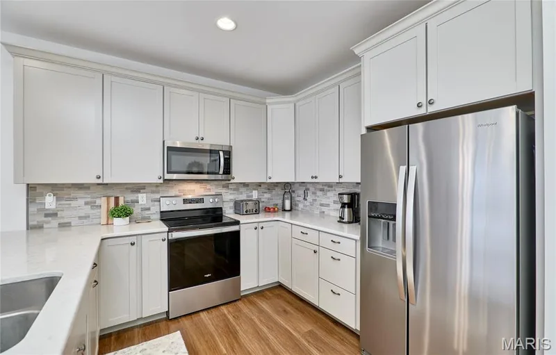 Kitchen featuring white upper and lower cabinetry, stainless steel appliances, light-toned countertops, tile backsplash, and wood-finish flooring — 305 Copperfield Court, St Charles, MO 63301