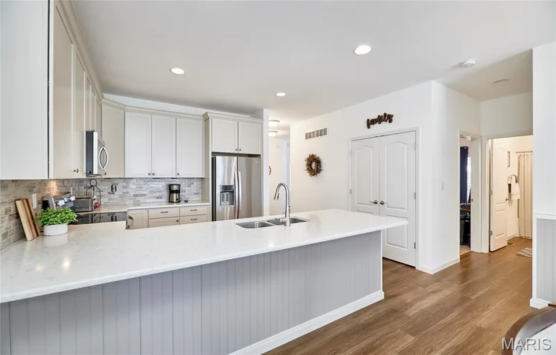 Open-concept kitchen featuring light-toned cabinetry, stainless steel appliances, a double basin sink, stone-finish backsplash, and wood-finish flooring — 305 Copperfield Court, St Charles, MO 63301