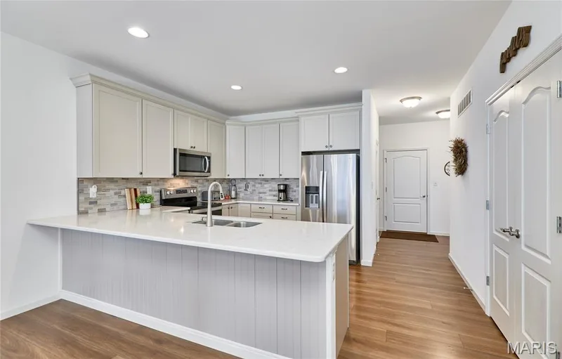 Kitchen featuring light-toned cabinetry, stainless steel appliances, a tile backsplash, a kitchen island with seating, and wood-finish flooring — 305 Copperfield Court, St Charles, MO 63301