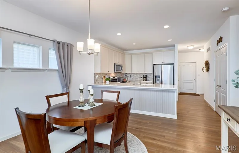 Open-concept dining area with wood-finish flooring, a kitchen featuring white cabinetry, stainless steel appliances, and a tile backsplash — 305 Copperfield Court, St Charles, MO 63301