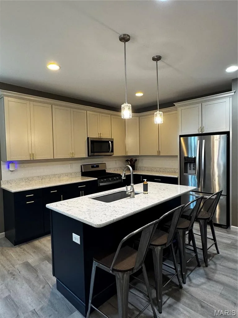 Well-appointed kitchen featuring light-toned upper cabinetry, dark lower cabinetry, stainless steel appliances, a center island with sink, and wood-finish flooring — 401 Copperfield Court, St Charles, MO 63301