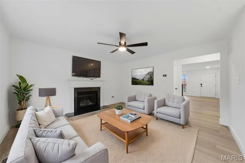 Living room featuring a fireplace with a white mantel, light wood-finish flooring, and an overhead ceiling fan — 8 Shady Elm Court, St Charles, MO 63301