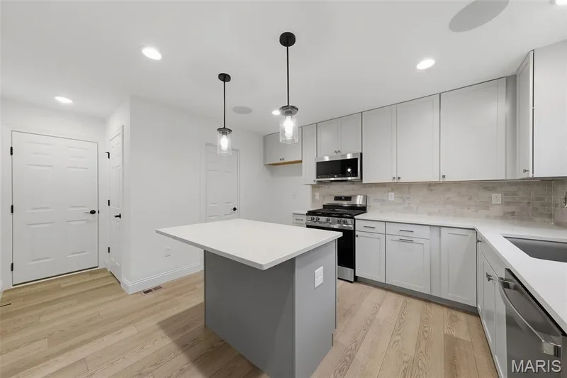 Kitchen featuring light wood-finish flooring, white cabinetry, integrated stainless steel appliances, a center island with pendant lighting, and a tile backsplash — 8 Shady Elm Court, St Charles, MO 63301