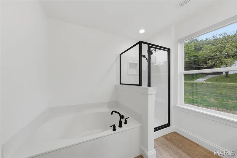 Corner soaking tub with matte black fixtures, adjacent to a glass-enclosed shower with black trim, wood-finish flooring, and a large window — 8 Shady Elm Court, St Charles, MO 63301