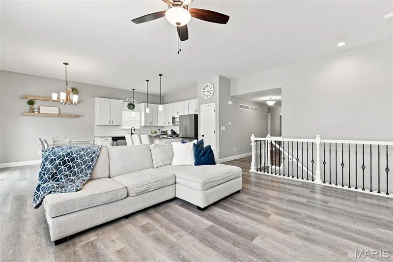 Open-concept living space featuring wood-finish flooring, a ceiling fan with integrated lighting, and a white spindle railing with black wrought iron balusters — 130 Pomodora Circle, Wentzville, MO 63385