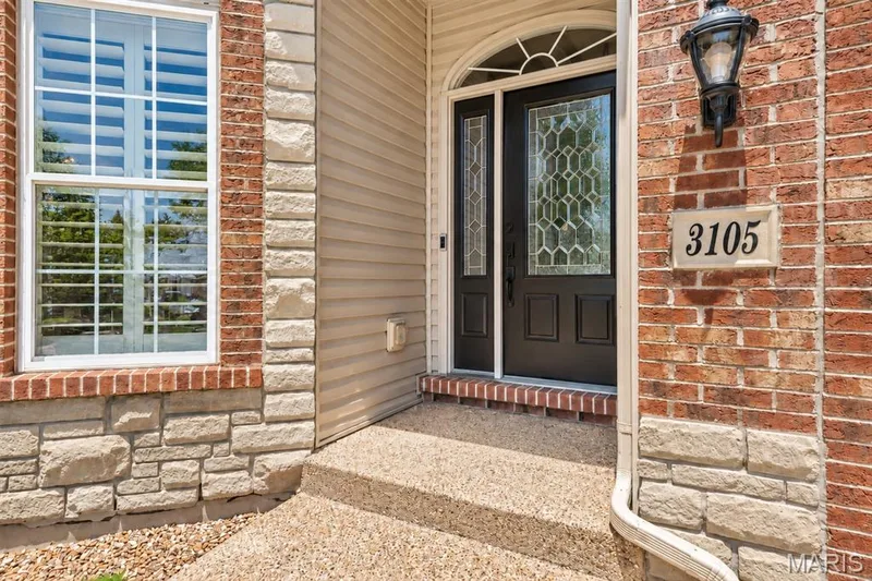 Brick and stone facade with an arched entry, decorative glass front door, and a wall-mounted exterior light fixture — 3105 Wheeling Drive, St Charles, MO 63301