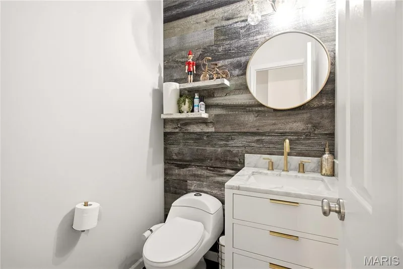 Wood-finish accent wall, white vanity with gold-tone hardware, undermount sink with gold-tone faucet, round wall mirror, and white wall shelving — 308 Graywynd Court, Lake St. Louis, MO 63367