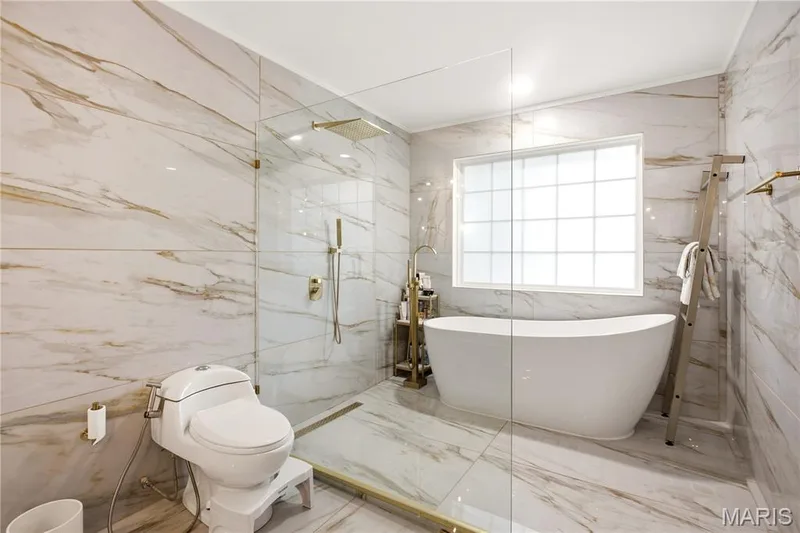 Full-height marble-look tiled walls and flooring with a freestanding soaking tub, enclosed glass shower, and gold-finish fixtures — 308 Graywynd Court, Lake St. Louis, MO 63367