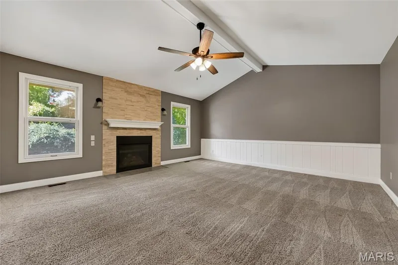 Vaulted ceiling with exposed beam, stone-clad fireplace with mantel, two windows, ceiling fan, and wainscoting — 19 Novella Drive, St Peters, MO 63376