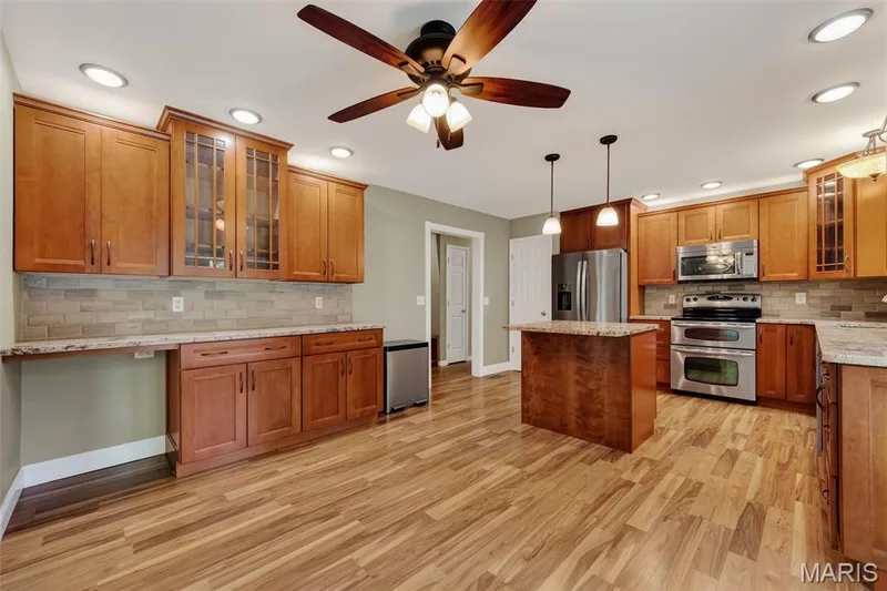 Kitchen featuring wood-finish flooring, wood cabinetry with some glass-front doors, a central island with a solid surface countertop, stainless steel appliances, and recessed lighting — 19 Novella Drive, St Peters, MO 63376