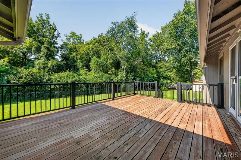 Expansive composite deck featuring warm wood-finish flooring and black metal railings, overlooking manicured lawn and mature tree line — 19 Novella Drive, St Peters, MO 63376