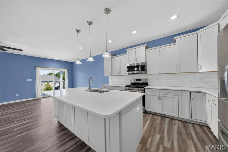 Kitchen featuring an expansive island with an undermount sink, white cabinetry, a subway tile backsplash, stainless steel appliances, and wood-finish flooring — 622 Cruden Bay Court, Dardenne Prairie, MO 63368