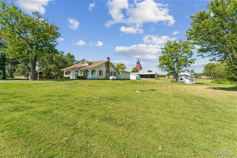 Expansive grassy property with multiple outbuildings, mature trees, and a house featuring a covered porch and brick chimney — 435 Highway T, Foristell, MO 63348