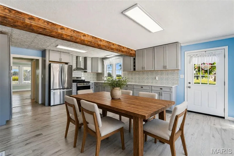 Expansive kitchen featuring light wood-finish flooring, light gray cabinetry, a tile backsplash, stainless steel appliances, and a wood beam accent — 435 Highway T, Foristell, MO 63348