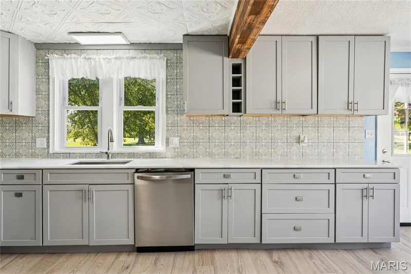 Light gray cabinetry with brushed nickel hardware, white countertop, patterned tile backsplash, stainless steel dishwasher, and wood-finish flooring — 435 Highway T, Foristell, MO 63348