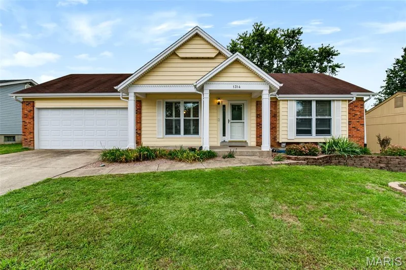 Exterior featuring yellow siding, brick accents, a two-car garage, and a covered porch entry — 1314 Warrior Song Way, O'Fallon, MO 63366
